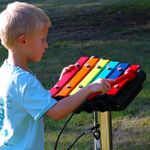 a young boy playing on a small rainbow coloured outdoor xylophone mounted on a stainless steel post in a playground