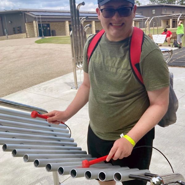 a young adult with special needs wearing a green t shirt and playing a large outdoor xylophone in a music park at summer camp 