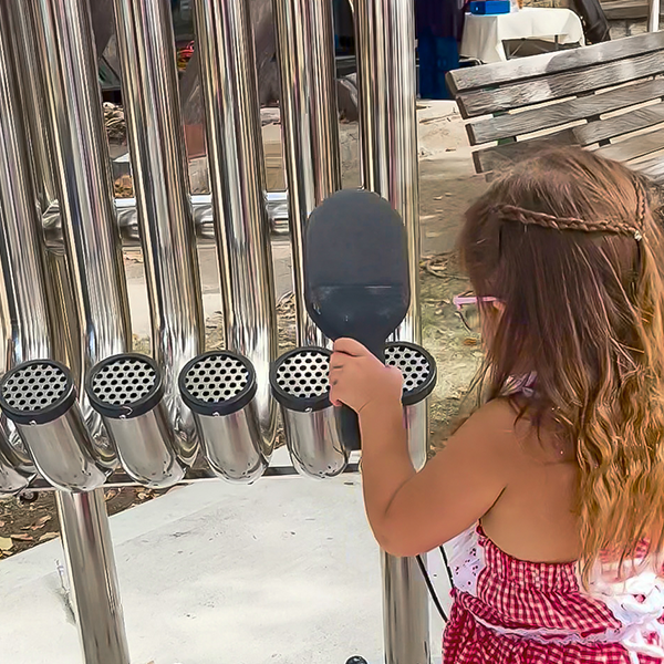 A young girl in a red and white dress plays outdoor pipes with a black mallet. The scene conveys curiosity and playfulness in a park setting.