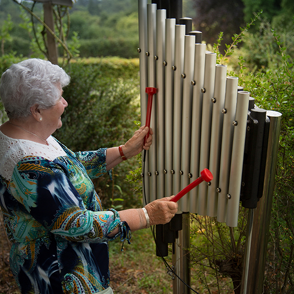 a senior lady playing an outdoor musical instrument that is vertically mounted and has 11 aluminium chimes. She is playing using red mallets.