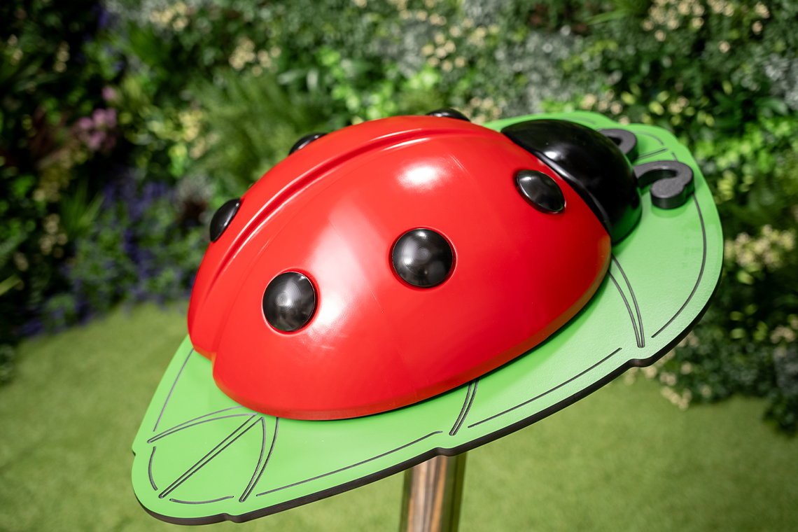 A large, glossy red ladybug musical sculpture with black spots sits on a green leaf platform indoors. Lush greenery forms the blurred background, creating a natural, playful ambiance.