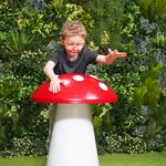 A young boy with glasses joyfully plays on a large red and white musical toadstool, surrounded by vibrant greenery, exuding a sense of wonder and playfulness.