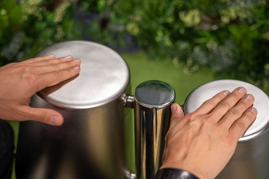 Two hands playing shiny steel drums outdoors, surrounded by lush green plants. The scene conveys a sense of harmony and relaxation.