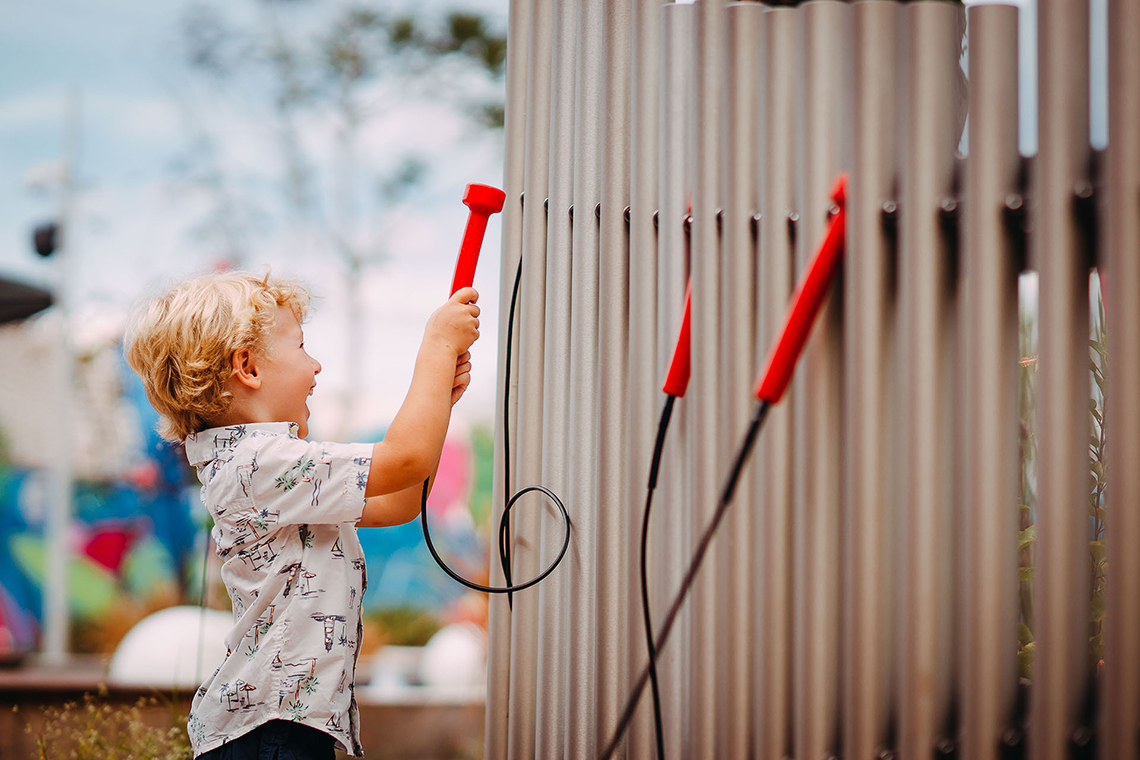 A blonde little boy playing a set of large musical chimes in a music park or playground