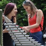 two young women laughing and playing a large outdoor musical instrument together in the park