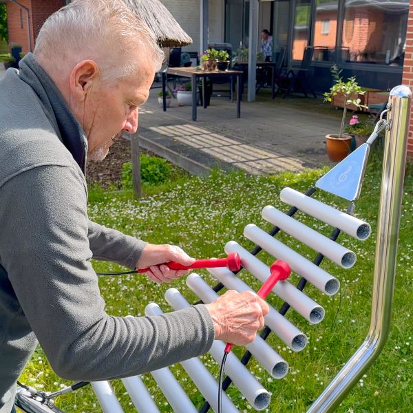 a senior man playing a large silver outdoor xylophone with red mallets 