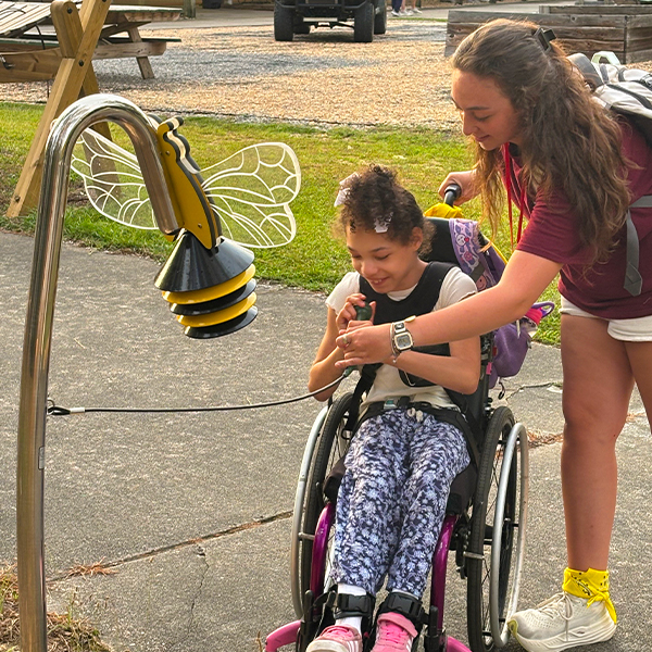 a young female wheelchair user and a camp counsellor playing an outdoor musical instrument shaped like a bumblebee in a camp playground