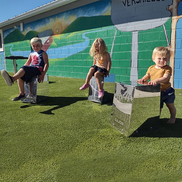 three young children sitting on and playing on three stainless steel cajon drums in the town of versailles music garden