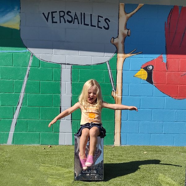 a young girl sitting on and playing a silver outdoor cajon drum at the town of versailles outdoor music garden