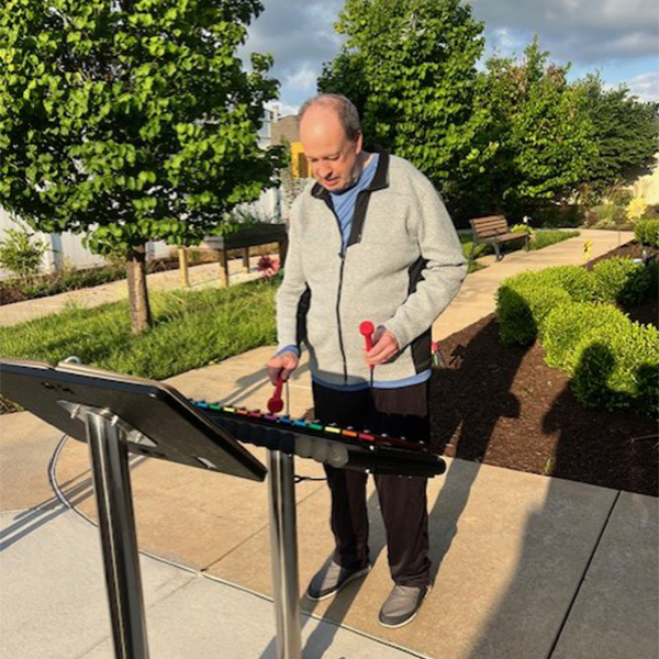 a senior man playing an outdoor musical xylophone with a music book on stand in front in a senior living garden