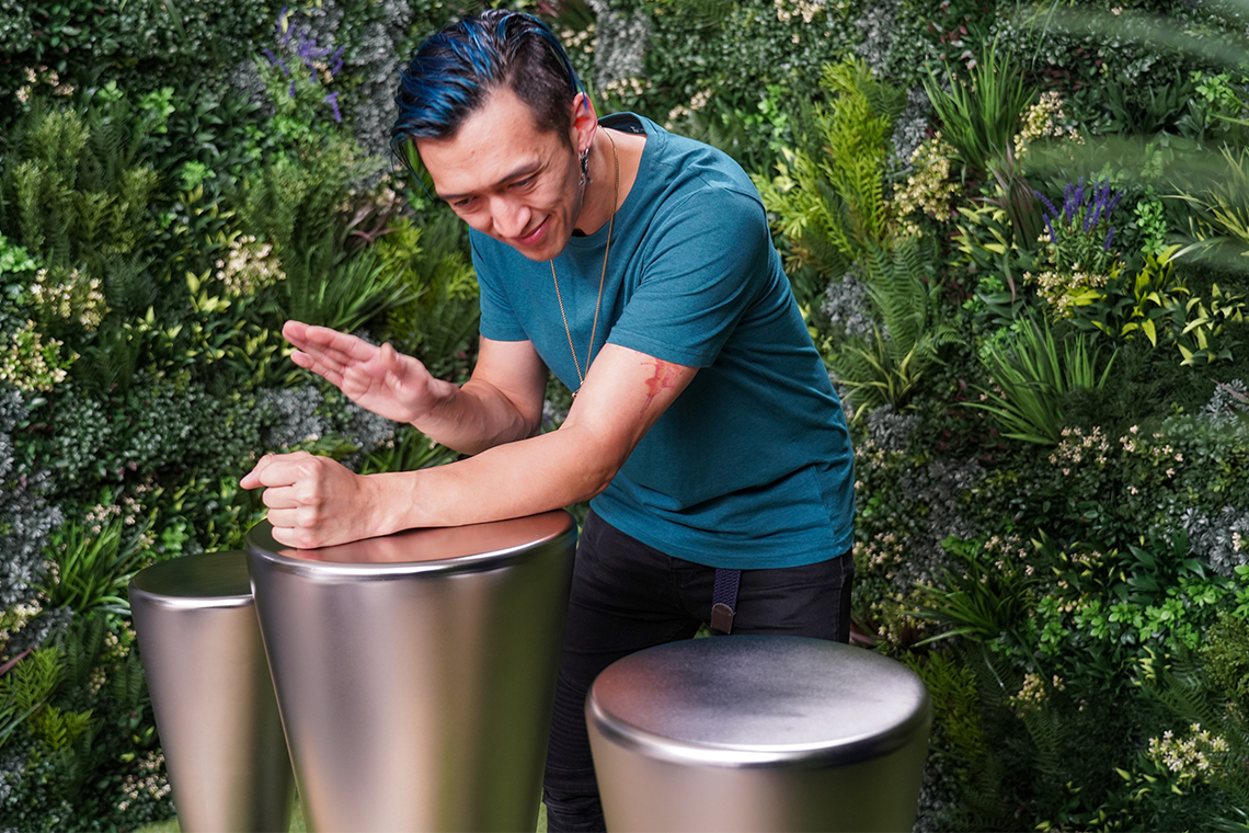 A person with blue hair smiles while playing sleek, metallic outdoor Djembe drum instruments in a lush, green garden setting, conveying joy and creativity.