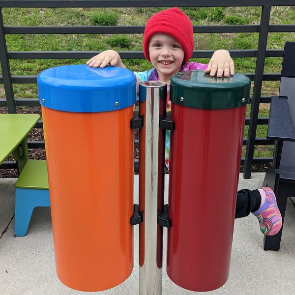 Young boy wearing a hat standing behind a pair of large colorful outdoor conga drums with his hands drumming the tops