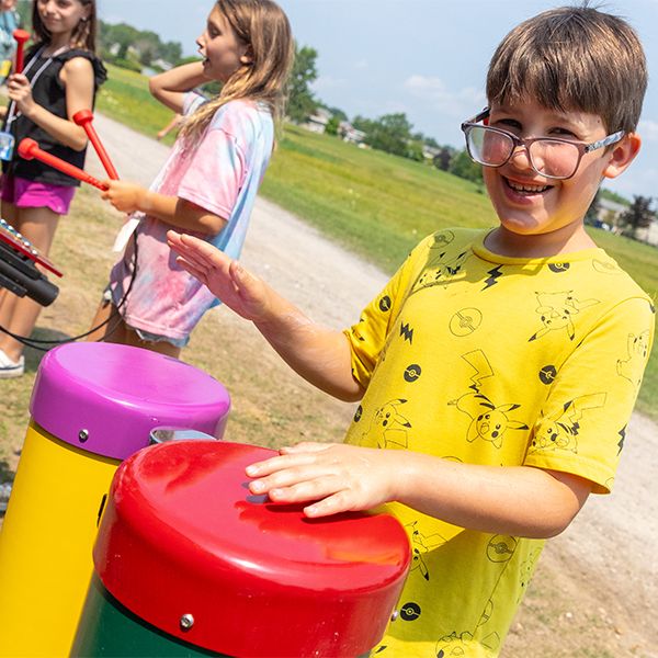 A boy wearing glasses and a yellow t shirt playing outdoor bongo drums