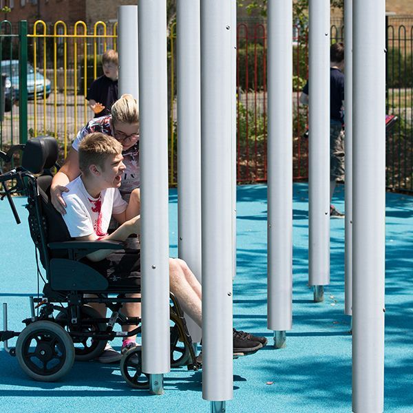 Young disabled man in a wheelchair with his carer playing on tall silver musical chimes in a school playground