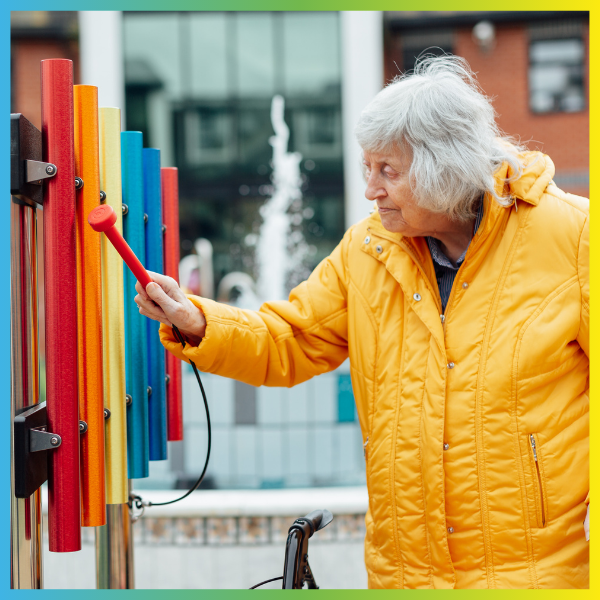 Senior lady in a yellow coat playing rainbow colored outdoor musical chimes in a music garden 