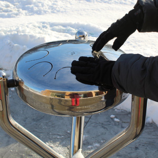 hands in black gloves playing a stainless steel tongue drum outdoors