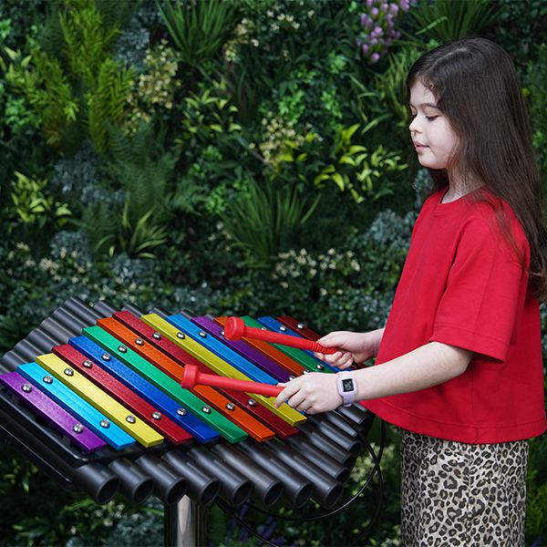 an outdoor xylophone with rainbow colored notes on a stainless steel leg with a young girl playing with red mallets and a green leafy background