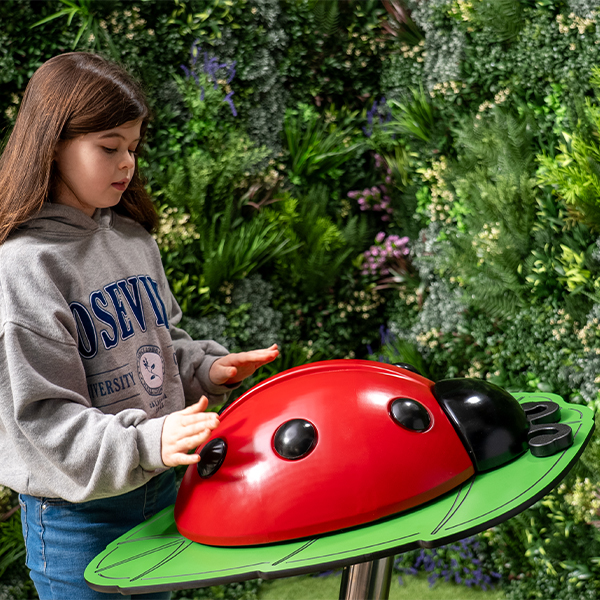 Young girl in a gray hoodie plays with a large, red ladybug musical sculpture on a green leaf platform. Background features lush greenery, creating a playful atmosphere.