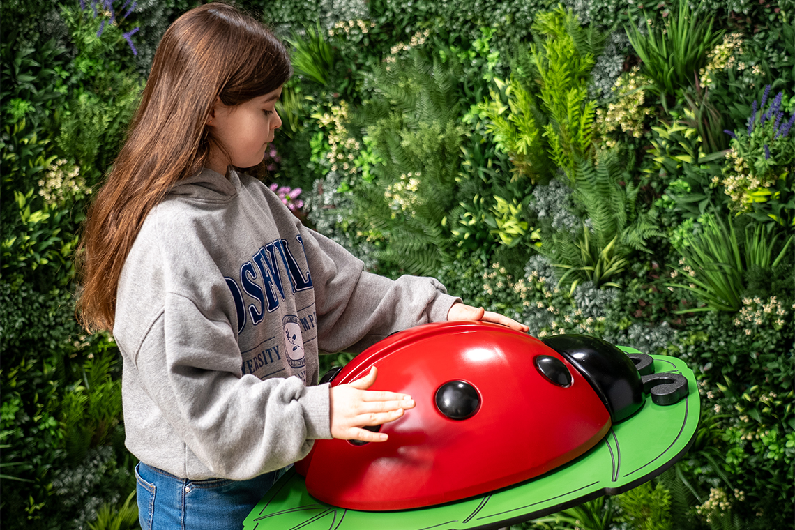 A girl in a gray hoodie playing a large musical ladybug on a green pedestal with her hands. The background is lush with green foliage, creating a playful, natural setting.