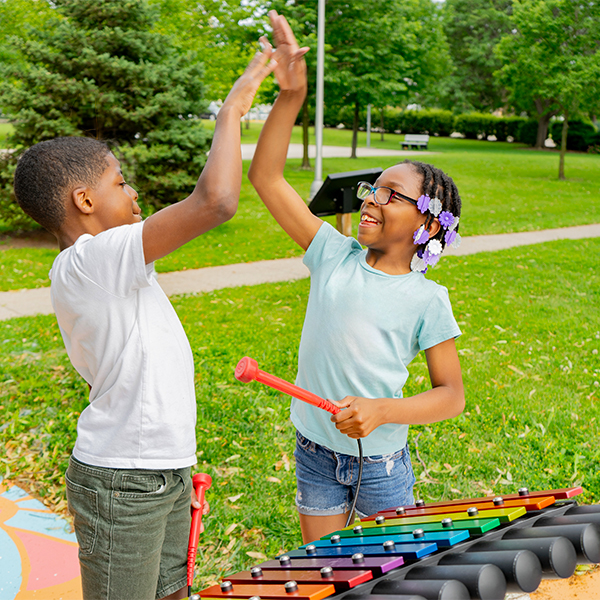 two young teens high fiving each other standing in front of an outdoor xylophone
