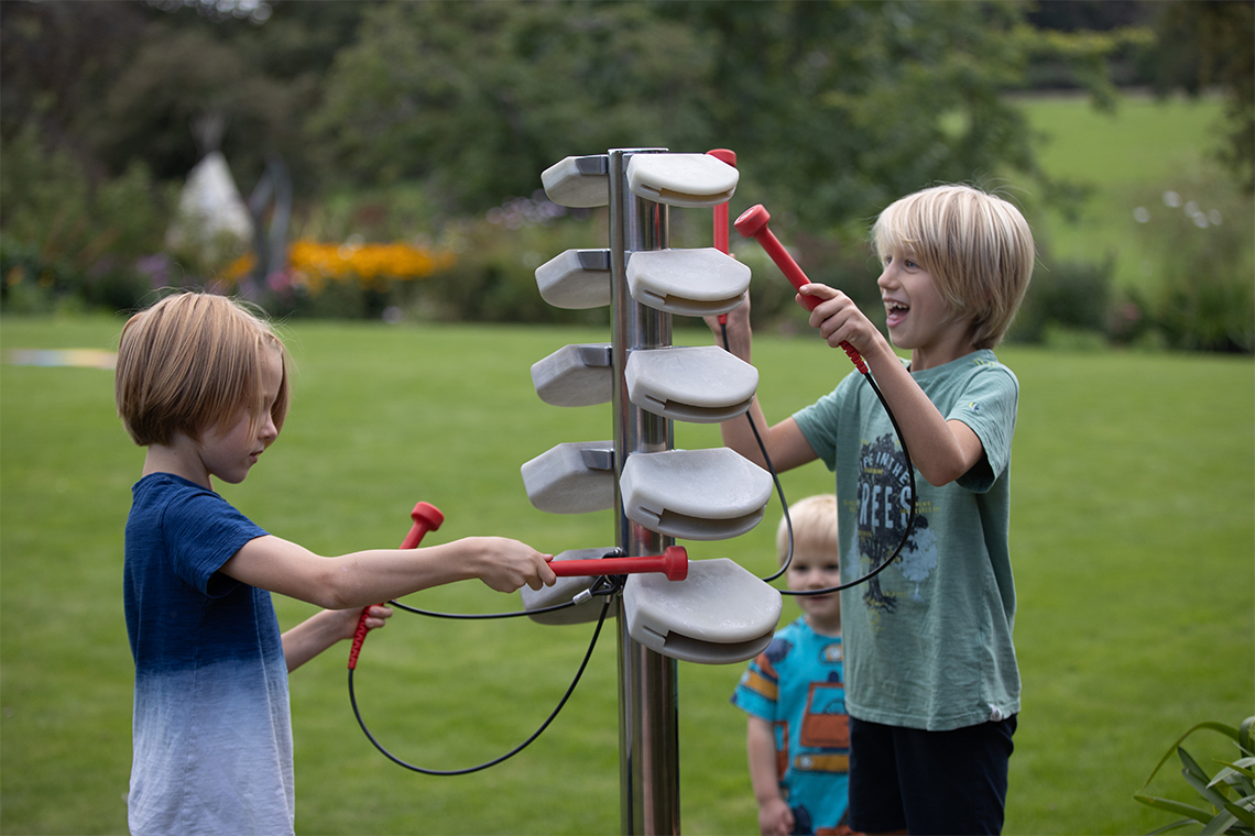 two boys playing an outdoor musical tree with branches made of stone colored temple blocks and with red mallets to hit them with