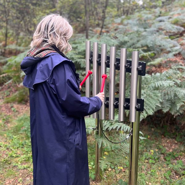 a lady in a long blue coat standing and playing a set of outdoor musical chimes in a woodland setting