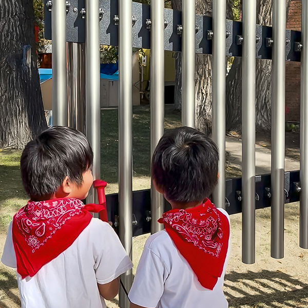 Two children wearing red bandanas play outdoor metal chimes. They stand side by side, focused, in a sunny park setting with trees in the background.