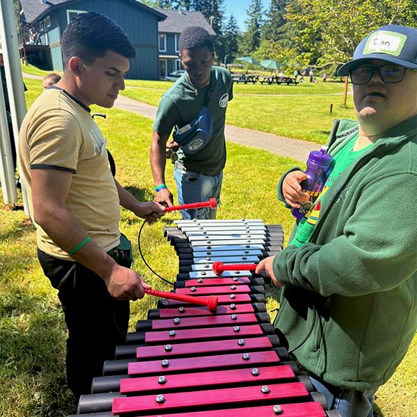 three young men, one with down's syndrome, playing an outdoor xylophone in the camp beausite new musical garden