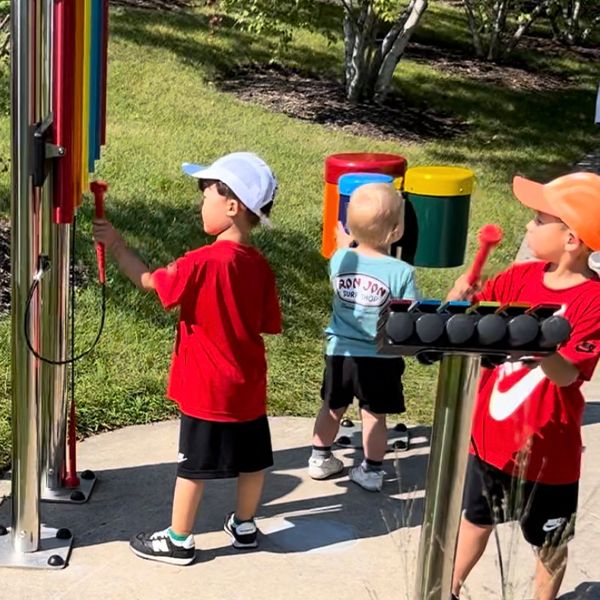 three young children wearing sun hats and red t shirts playing outdoor musical instruments in the Kohl museum garden