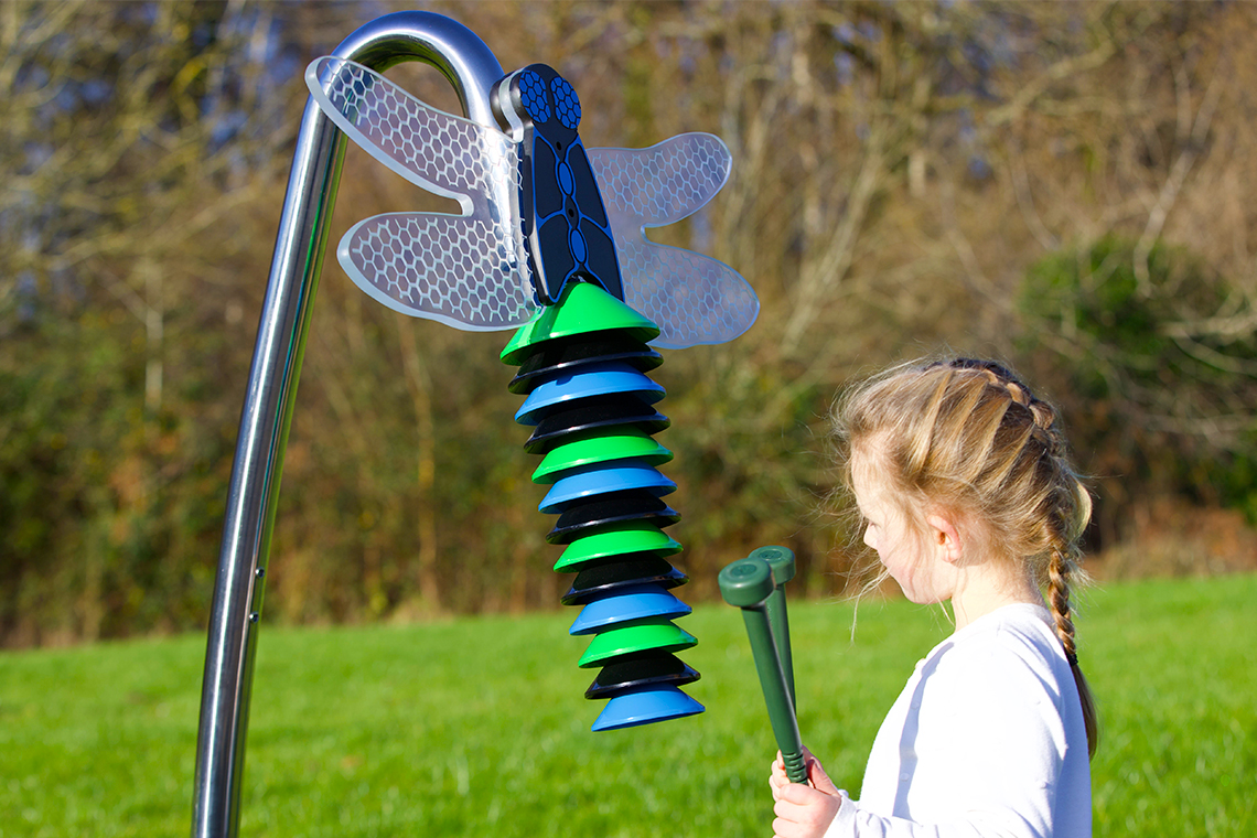 A child with braided hair plays with an outdoor musical instrument resembling a dragonfly. The bright green and blue discs stand out against a grassy background.