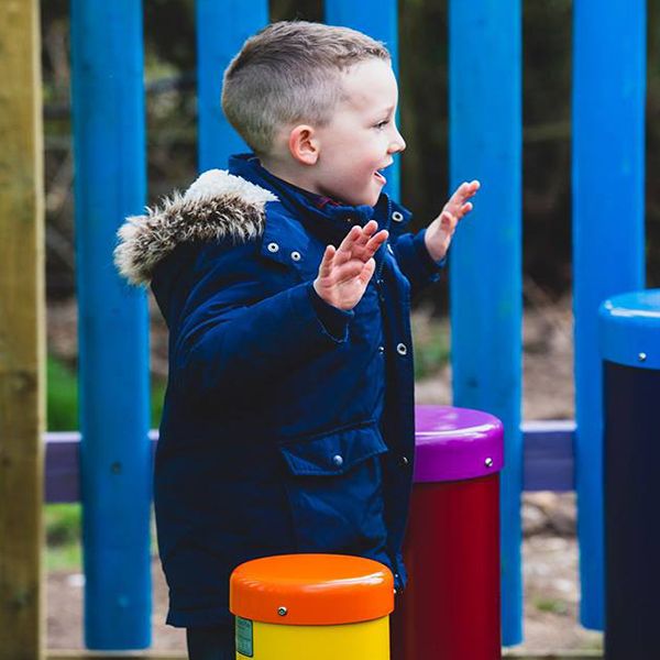 a young boy in a winter coat in a music playground playing two colourful outdoor drums with his hands