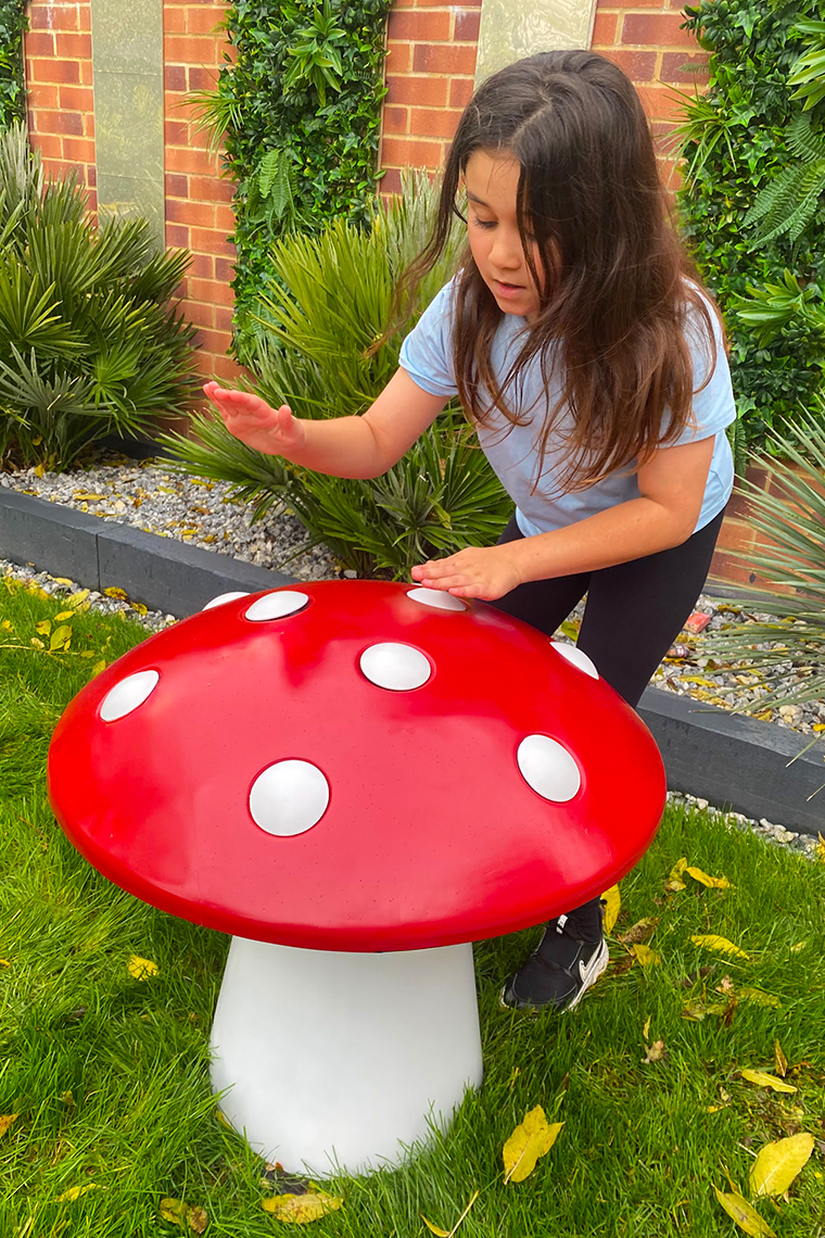 A girl in a blue shirt playfully interacts with a large, red mushroom musical sculpture with white spots in a garden. The scene is vibrant and whimsical.