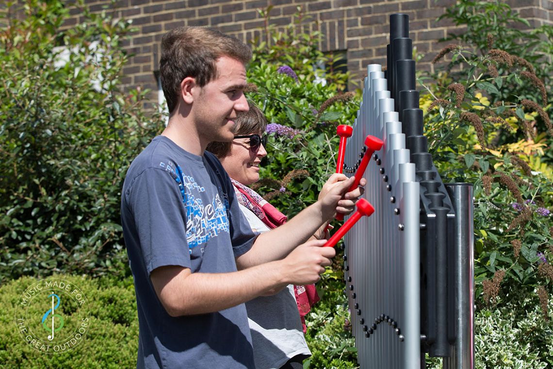 two people playing large outdoor musical instrument with upright tubular notes mounted on black resonators