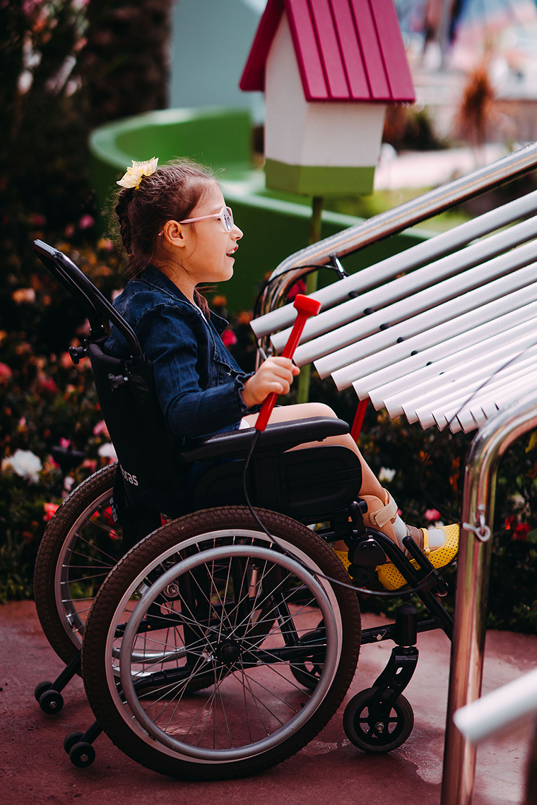 a young girl with special needs and in a wheelchair smiling and playing a large metal outdoor musical instrument in a playground