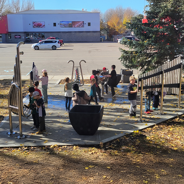 A group of children play with outdoor musical instruments at a playground, with a parking lot and trees in the background.