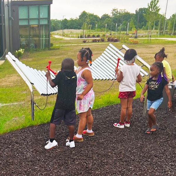 A group of young children playing a large silver outdoor musical instrument at Dayton Metro Library