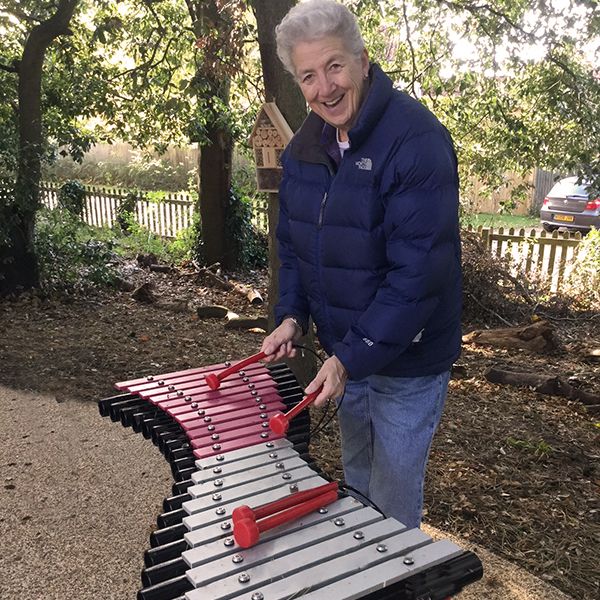 lady with grey hair playing a pink and silver large outdoor xylophone in the Aldeburgh hospital Sensory Garden