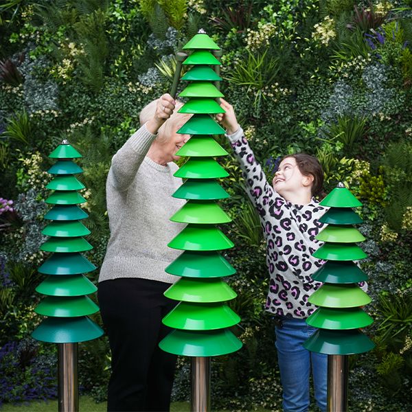 A child and elderly person smile while playing on green, bell sculptures resembling trees in front of a lush, leafy wall. The mood is joyful and collaborative.