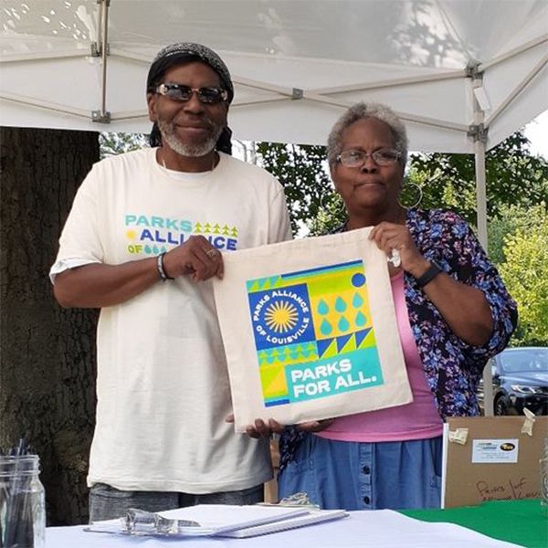 two older adults holding up a tote bag that says PArks for All and the Parks Alliance of Louisville logo