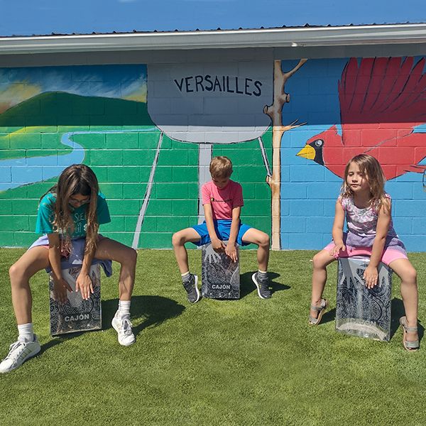 three young children sitting on and playing on three stainless steel cajon drums in the town of versailles music garden