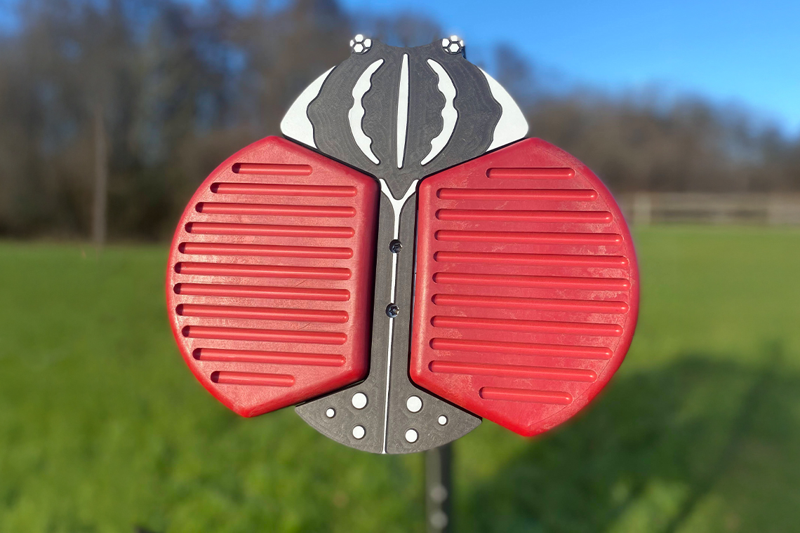 A vivid red and black bug-themed percussion instrument stands against a blurred green field and blue sky. The red wings have textured surfaces.