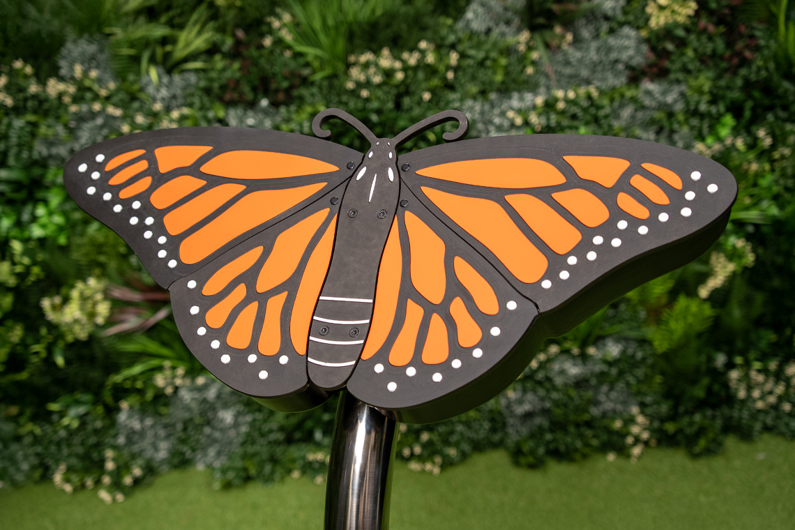 A large, monarch butterfly musical sculpture featuring orange wings with black outlines and white spots, set against a green foliage backdrop.