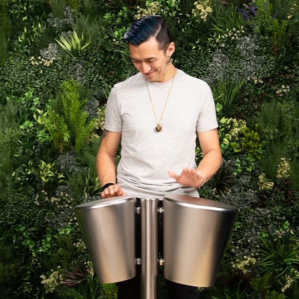 A person in a gray shirt energetically plays two large stainless steel drums. They are surrounded by a lush vertical garden, exuding a lively and joyous vibe.