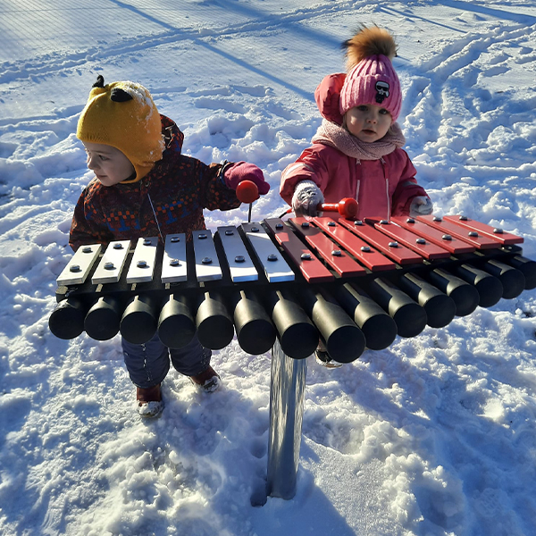 two young children dressed for cold snowy weather playing an outdoor xylophone together in a park