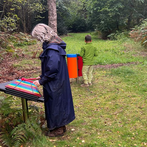 a lady wearing a long blue coat playing an outdoor rainbow colored xylophone with a small child playing bongs in the background in a woodland setting