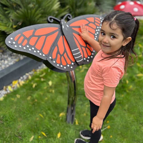 A young girl smiles while touching a large, colorful butterfly musical sculpture with orange and black wings in a garden, conveying joy and curiosity.