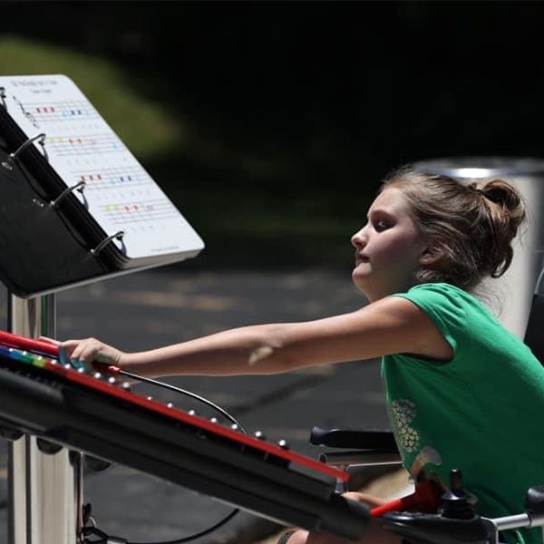 a young teenage girl in a wheelchair playing an outdoor xylophone in a music park at summer camp