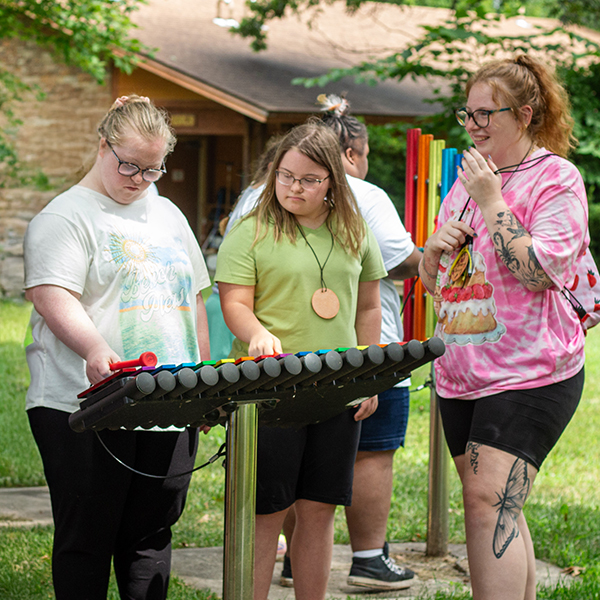 Three teenage girls standing and playing an outdoor xylophone in a percussion park at summer camp