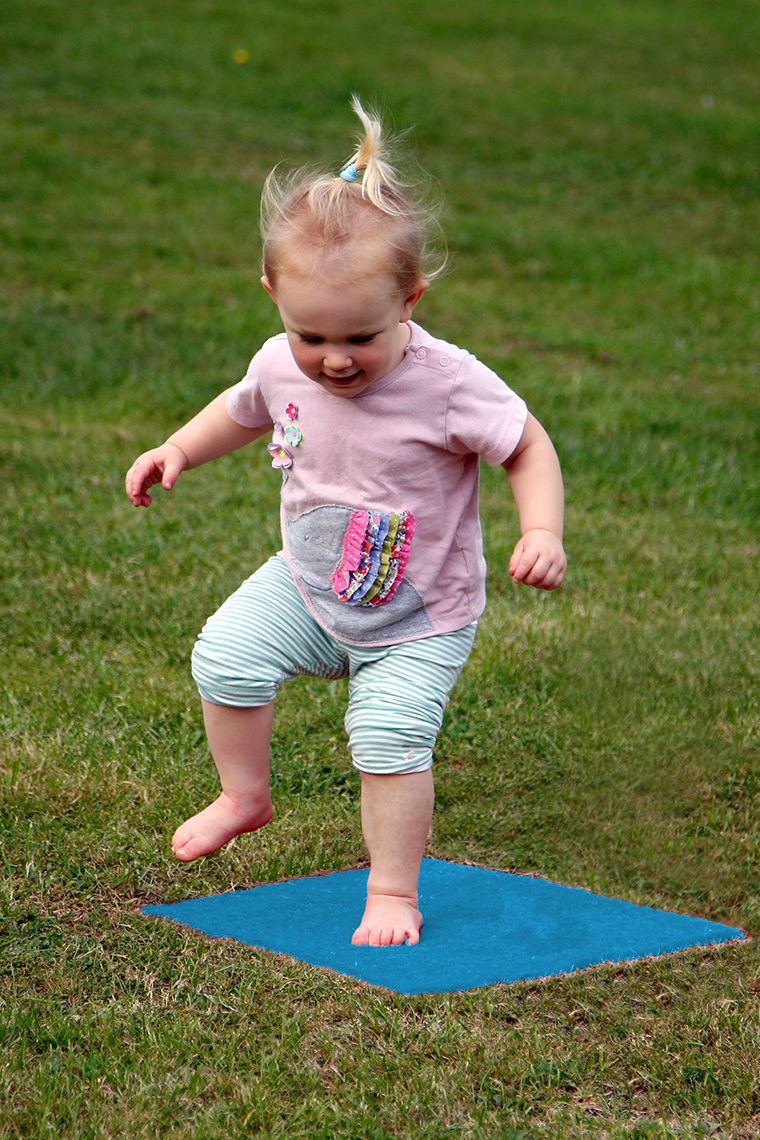 a young girl jumping on a blue musical stepping stone square in a lawn which is disguising musical notes