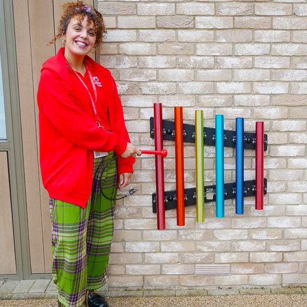 A lady in red jacket and green checkered trousers standing next to a set of rainbow colored chimes mounted on the wall in a childrens hospice